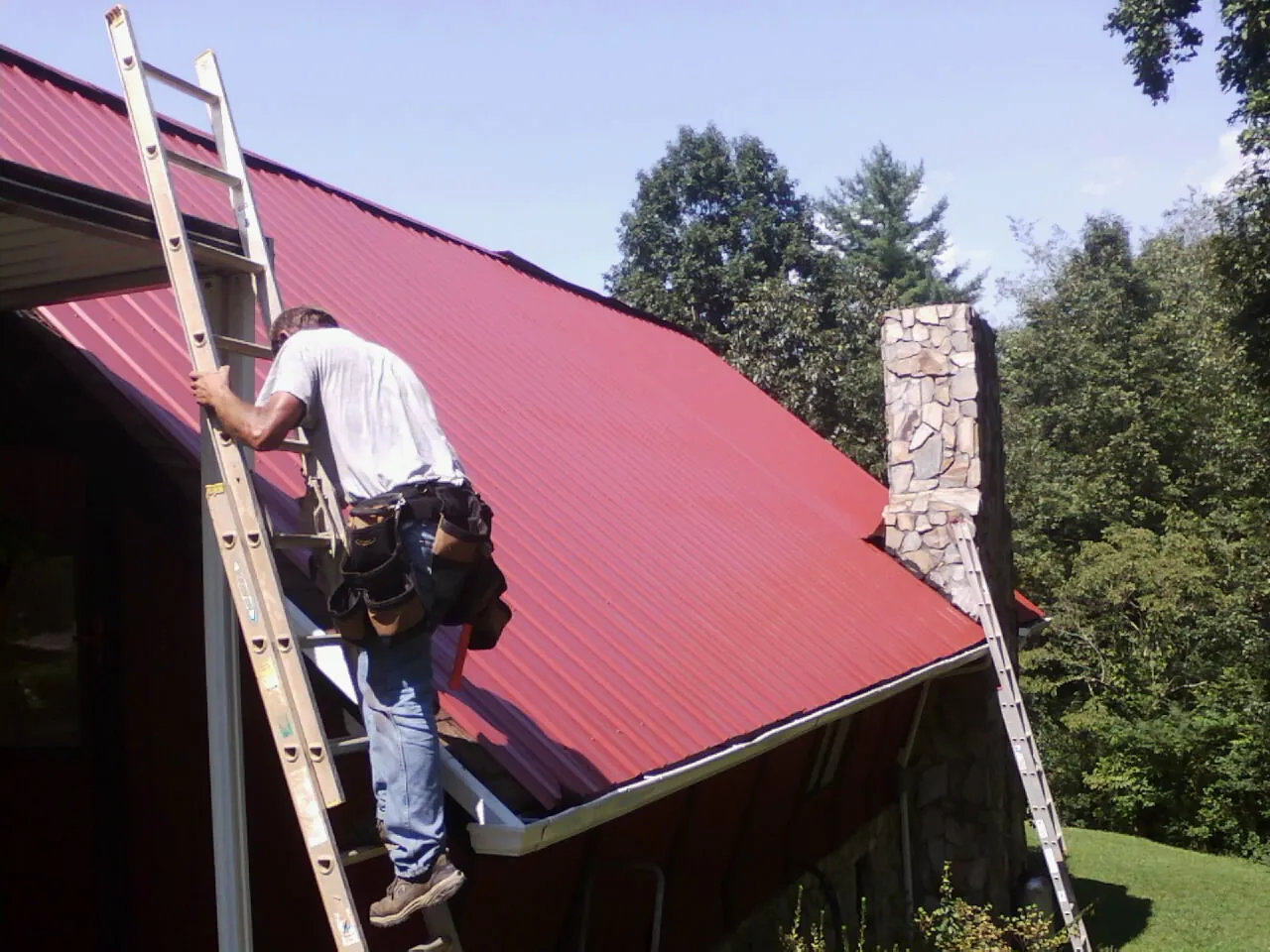 Roofer on a ladder beside a red metal roof and stone chimney — Soffit & Fascia Repair by Roofing Brothers in Prineville